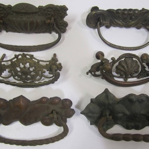 May include: Six antique bronze drawer pulls with ornate designs. The pulls feature various shapes, including floral and geometric patterns, with curved handles. The bronze finish gives them a vintage look, suitable for furniture restoration or decorative projects.