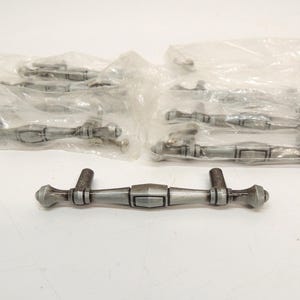 May include: A collection of silver-toned metal drawer pulls with a brushed finish. Each handle features a central rectangular block, tapered arms, and rounded end caps. Several of these hardware pieces are individually wrapped in clear plastic.