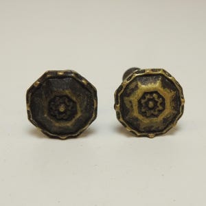 May include: Two round cabinet knobs with an octagonal outer edge and a dark, weathered brass finish. Each knob features a central raised floral or star-shaped motif surrounded by a textured, darkened surface and a highlighted brass rim.