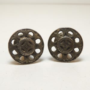 May include: A pair of circular silver-toned metal cufflinks featuring an openwork design with a central cross motif surrounded by eight small cutouts. The metal has a weathered, dark patina finish with visible texture and raised edges.