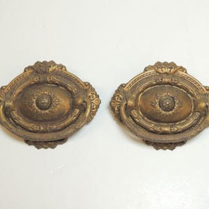 May include: Two oval-shaped brass drawer pulls featuring an ornate, raised relief pattern with a central rosette and a swinging bail handle. The metal has a darkened, aged patina with intricate scrollwork detailing along the outer edges.