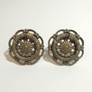May include: Two circular brass drawer pulls featuring an ornate, openwork outer frame and a central floral medallion. Each piece includes a hinged, wreath-shaped drop handle that rests against the decorative backplate, showcasing a dark patina.
