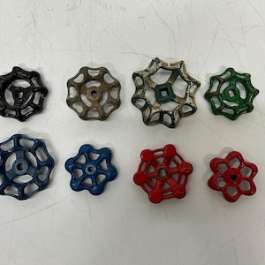 May include: An assortment of eight vintage water valve handles in various colors. The handles are in black, brown, green, blue, and red. The handles have a circular design with a star-like shape and are made of metal.