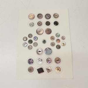 May include: A collection of assorted buttons arranged on a white display card. The buttons vary in size, shape, and color, featuring shades of gray, brown, iridescent pearl, and black. Some are circular, while others are square or diamond-shaped.