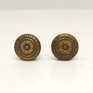 May include: Two circular brass cabinet knobs featuring a central raised floral motif surrounded by a textured border with repeating geometric patterns. The metal has a muted, aged gold finish with visible detailing on the petals and outer rim.