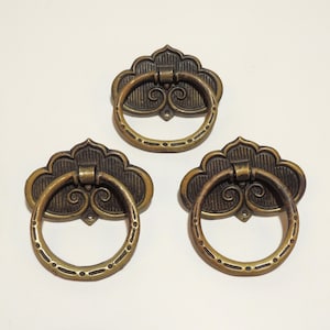 May include: Three antique brass-toned metal drawer pulls, each featuring a circular ring handle attached to a decorative, fan-shaped backplate with embossed linear detailing and a central scroll motif, arranged in a triangular formation.