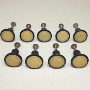 May include: A set of nine circular cabinet knobs featuring a cream-colored center disc surrounded by a dark brown rim. Each knob has a metallic threaded screw extending from the back, designed for attachment to furniture or cabinetry doors.