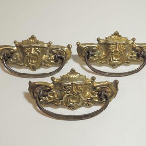 May include: Three brass drawer pulls featuring a detailed relief of a face with an open mouth. Each pull has a curved, textured metal handle attached to the ornate backplate. The brass has a warm, metallic gold finish with dark patina accents.