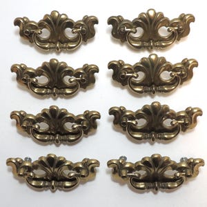 May include: A set of eight brass-toned metal drawer pulls arranged in two columns. Each pull features a decorative, ornate backplate with a curved, swinging handle attached to two mounting points, showcasing a polished metallic finish.