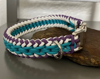 Puede incluir: Un collar de perro trenzado en color turquesa, blanco y morado con una hebilla y un anillo en D plateados.