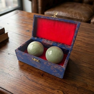 May include: A blue wooden box with a red velvet interior holds two smooth, pale green spheres. The box has a gold clasp and is open, revealing the contents. The spheres appear to be about 2 inches in diameter.