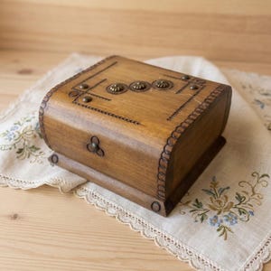 Op de afbeelding: Een rechthoekige houten doos met een gebogen deksel, voorzien van een medium bruine afwerking. Het deksel is versierd met drie kleine ronde metalen noppen op een rij, omringd door geëtste lijnpatronen en een geschulpte rand.