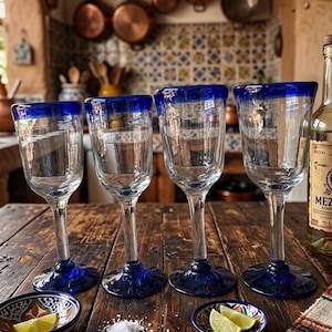 Könnte beinhalten: Vier transparente Weingläser mit einem leuchtend blauen Rand und Boden stehen auf einem rustikalen Holztisch. Kleine Schalen mit Limettenspalten und Salz befinden sich im Vordergrund. Eine Flasche Mezcal ist rechts zu sehen.
