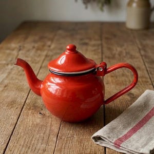 May include: A red enamel teapot with a curved handle and spout. The teapot has a lid and sits on a wooden surface. A folded linen towel with red stripes is next to the teapot.
