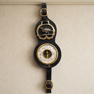 Op de afbeelding: Een wandbarometer bevestigd in een zwarte leren riem met koperen gespen. Het bovenste gedeelte bevat een koperen vogel-embleem, terwijl het onderste ronde gedeelte een witte wijzerplaat heeft met zwarte tekst zoals Stormy, Rain, Change, Fair en Set Fair.