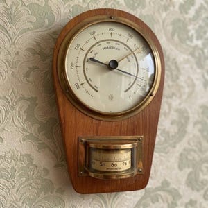 May include: A wall-mounted weather station featuring a circular brass-rimmed barometer with a cream-colored dial and black needles, mounted on a teardrop-shaped wooden plaque. Below the barometer is a smaller brass-encased hygrometer with a gauge.