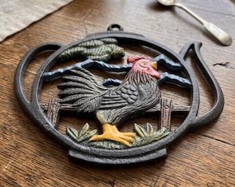 May include: A cast iron trivet in the shape of a teapot, featuring a rooster design. The rooster has red, black, and yellow coloring, with a fence and foliage in the background. The trivet is on a wooden surface.