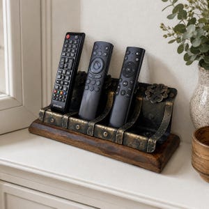 May include: A wooden remote control holder with three compartments, each holding a black remote. The holder has a dark brown base and is accented with brass-colored metal. The holder is on a white surface.