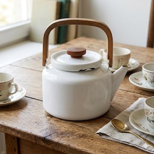 May include: A white enamel teapot with a rounded body, a matching lid featuring a small brown wooden knob, and a curved wooden handle. The teapot has a short spout and is positioned among several small ceramic teacups on saucers.