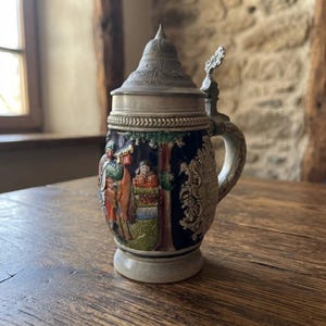 Peut inclure: Une chope à bière en céramique avec un corps bleu foncé orné d'un relief coloré représentant un personnage avec un cor, un paysage et des volutes. Elle comprend un couvercle métallique avec charnière et une anse incurvée sur le côté.