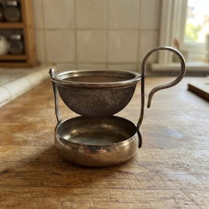 May include: A silver-toned metal tea strainer with a fine mesh bowl suspended above a matching circular drip tray. The strainer features a curved handle extending from the side, connecting the upper mesh component to the solid base below.