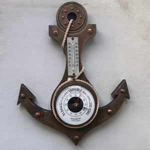 May include: A dark brown anchor-shaped wall instrument featuring a vertical thermometer with Celsius markings and a round barometer dial. The barometer face displays the French text Tempête, Variable, and Très-Sec, with a rope accent.