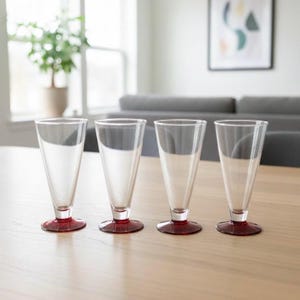 May include: Four clear stemmed glasses with a flared design and a red base are displayed on a light wooden table. The glasses are empty. The background features a window, a potted plant, and a grey sofa. The glasses are suitable for cocktails.