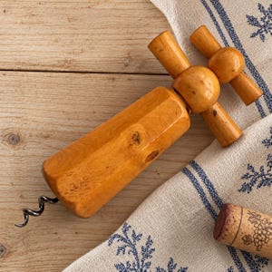 Peut inclure: Un tire-bouchon en bois avec une finition polie brun clair et un grain de bois apparent. L'outil présente une poignée en forme de T et une mèche en métal en spirale. Un bouchon cylindrique avec une illustration de vigne est posé à côté.