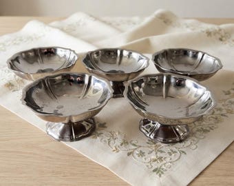 May include: A set of four silver-toned metal dessert bowls, each featuring a scalloped rim, a smooth interior basin, and a sturdy pedestal base. The bowls have a reflective, polished finish and are arranged in a cluster on a flat surface.
