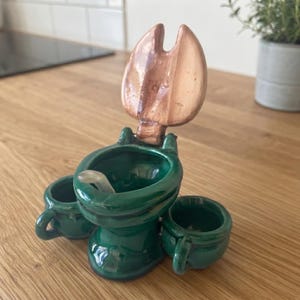 May include: A dark green ceramic toilet-shaped vessel featuring a glossy finish, a raised copper-colored lid, and two small matching green mugs attached to the sides. A small white spoon rests inside the central bowl of the ceramic structure.