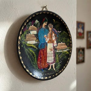 May include: A round black decorative wall plate featuring a hand-painted scene of two figures in traditional folk attire standing in a landscape with thatched-roof houses. The rim is decorated with gold leaf patterns and Cyrillic text at the bottom.