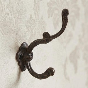 Könnte beinhalten: Ein dunkelbrauner Wandhaken aus Metall mit einem zweizinkigen Design und einer verzierten, geschwungenen Silhouette. Die Basis ist mit zwei sichtbaren Schrauben befestigt und die Metalloberfläche hat ein texturiertes, gealtertes Finish.