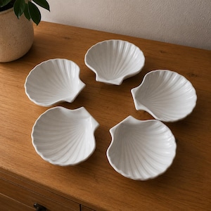 Peut inclure: Un ensemble de cinq coupelles en céramique blanche en forme de coquilles Saint-Jacques. Chaque coupelle présente une finition brillante et une série de stries rayonnantes partant de la charnière vers le bord festonné, créant un aspect uniforme.
