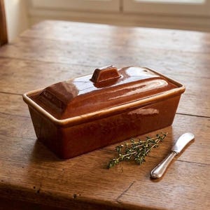 May include: A rectangular, brown ceramic butter dish with a lid. The dish is on a wooden surface, next to a sprig of thyme and a small silver knife. The dish has a glossy finish and a small handle on the lid.