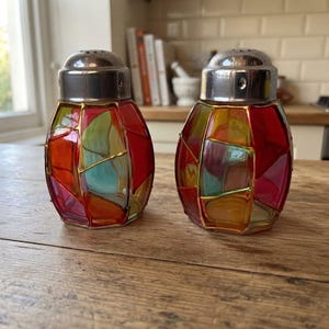 Puede incluir: Dos saleros y pimenteros con un diseño de vidriera con paneles multicolores en rojo, amarillo y azul, separados por líneas de metal dorado. Cada recipiente tiene un cuerpo de vidrio redondeado y una tapa de metal plateado pulido en la parte superior.