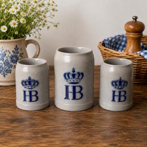 Peut inclure: Trois chopes à bière en grès gris ornées d'un logo couronne bleu et des lettres HB. Les chopes ont une forme cylindrique et une finition texturée. Le motif est identique sur les trois pièces, avec le logo centré sur le devant de chaque chope.