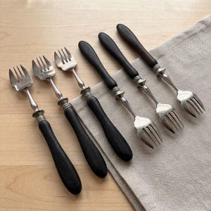 May include: A set of six small forks with dark matte black handles and silver-toned metal tines. Each fork features a rounded handle design with a ribbed metal collar connecting the handle to the fork head. The forks are arranged in a row.