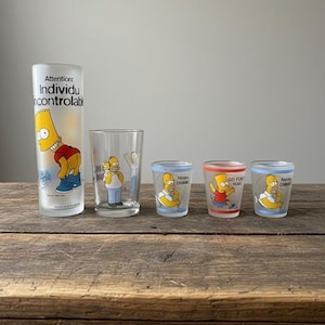 May include: A set of Simpsons-themed drinking glasses. The set includes a tall glass with Bart Simpson, a medium glass with Homer Simpson, and three shot glasses. The shot glasses feature Bart and Homer with phrases.