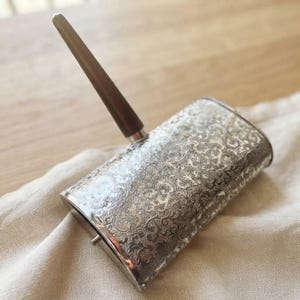 May include: A rectangular silver-toned metal desk accessory featuring an intricate, embossed floral scrollwork pattern across its surface. A single, smooth, dark brown wooden pen or stylus is inserted into a circular holder on the top edge.