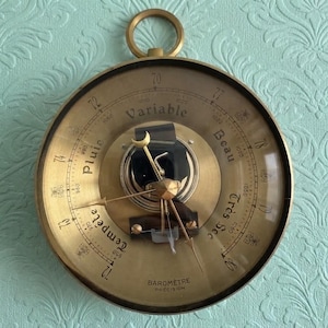 May include: A round brass barometer with a glass face featuring a central needle and a crescent moon pointer. The dial displays French weather terms including Pluie, Variable, Beau, Très Sec, and Tempête, along with numerical pressure scales.