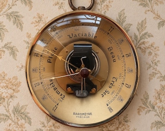 May include: A round brass barometer with a glass face featuring a gold-toned dial. The dial displays numerical scales, the text Variable, Pluie, Beau, Tempête, Très Sec, and Baromètre Précision, with thin metal needles and a central mechanism.