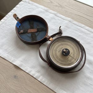 Könnte beinhalten: Ein runder, brauner Lederbehälter mit Klappdeckel, der ein beigefarbenes Instrument mit schwarzen Markierungen und der Aufschrift "Yvon Paris" enthält. Die Innenseite des Deckels ist mit blauem Stoff ausgekleidet und hat einen Metallriemen.