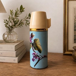 Può includere: Termos vintage con corpo azzurro e coperchio beige. Il thermos presenta un motivo floreale con fiori viola e foglie verdi. Una fascia dorata accentua la parte superiore del corpo blu. Il thermos è esposto su una superficie di legno.