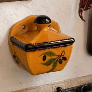 Op de afbeelding: Een aan de muur gemonteerde zoutbak van keramiek met een glanzende mosterdgele afwerking en een donkerbruin deksel. De voorzijde is voorzien van een handgeschilderd groen olijftakontwerp met zwarte olijven. Het deksel heeft een ronde knop.