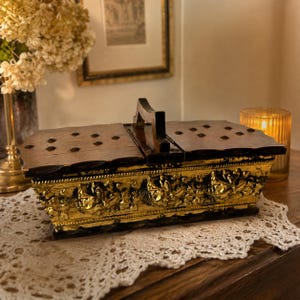 Pode incluir: Uma caixa de madeira vintage com tampa articulada e alça. A caixa apresenta um design dourado e ornamentado ao redor da base e um acabamento em madeira escura na tampa. A caixa está sobre uma toalha de renda branca.