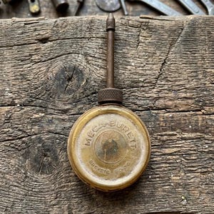 Op de afbeelding: Een vintage ronde metalen oliekan met een lange, dunne tuit en een gekartelde dop. De behuizing bevat de in reliëf aangebrachte tekst MECA-BURETT en BREVETE S.G.D.G. Het oppervlak heeft een verweerde, doffe messing afwerking met een textuur in het midden.