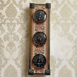 Könnte beinhalten: Eine vertikale Wetterstation zur Wandmontage mit einem rechteckigen Rahmen und einer floralen Gobelin-Einlage. Sie enthält drei runde, schwarze Messgeräte mit weißen Markierungen, darunter ein Barometer, ein Hygrometer und ein Thermometer in Messing.
