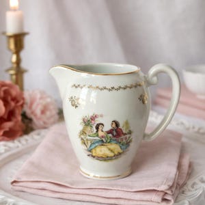Peut inclure: Une crémière en porcelaine blanche avec un bord et une anse dorés. La crémière présente une scène peinte d'un couple dans un jardin, avec des accents floraux et dorés. Elle repose sur une serviette en lin rose.