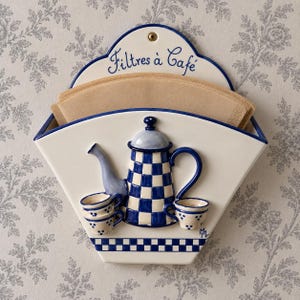 Può includere: Un portafiltri da caffè in ceramica color crema da parete con una teiera a scacchi blu e bianchi e tre tazzine. La parte superiore presenta la scritta Filtres à Café in blu. Contiene una pila di filtri di carta marrone.