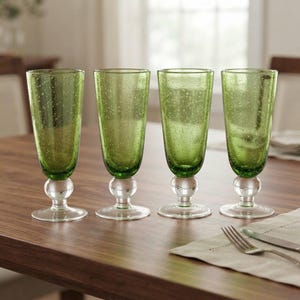 May include: A set of four tall green drinking glasses with a textured bubble finish. Each glass features a clear, spherical glass accent on the stem and a circular base. The glasses are arranged in a row, showcasing the consistent green hue and glass texture.